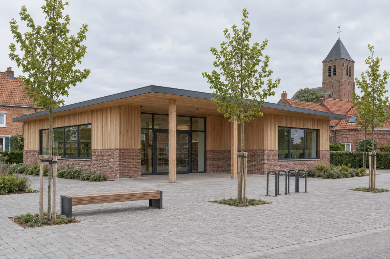 Bibliothèque et salle des associations dans un village des Weppes : soubassement en brique de réemploi, bardage en mélèze, auvent sur poteaux de bois et parvis planté, clocher de brique en arrière-plan