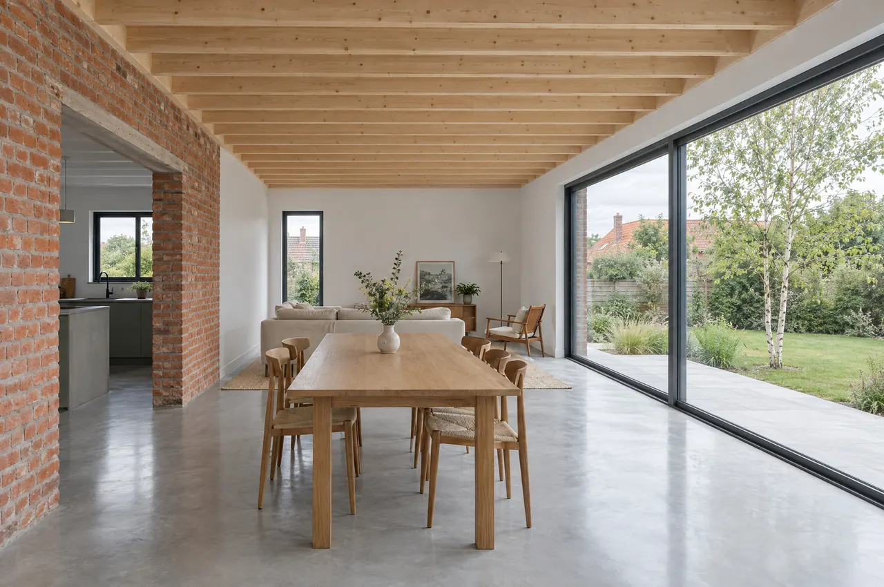 Intérieur de l’extension : plafond en solives d’épicéa, sol en béton poli, table en chêne et grande baie vitrée sur le jardin, ouverture dans le mur de brique vers la cuisine